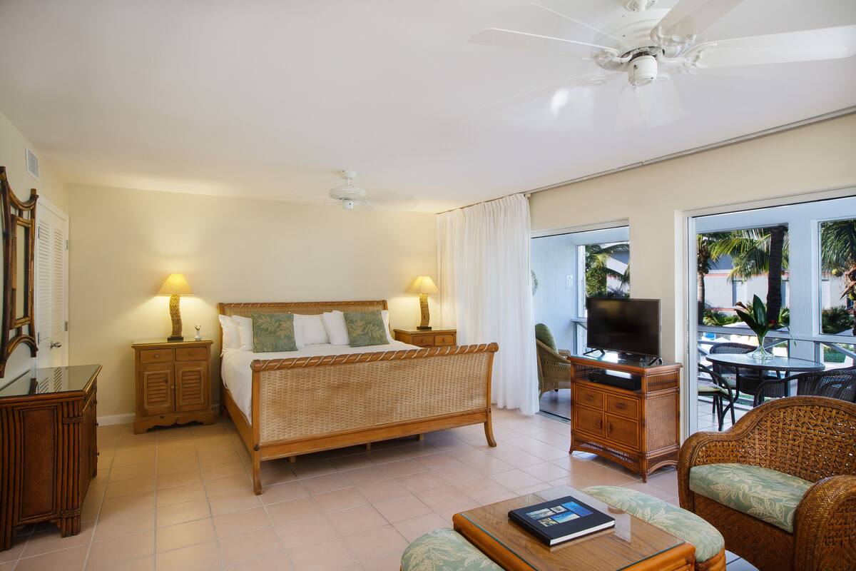 Quarto de hotel com cama, mesas de cabeceira, televisão, área de refeições com mesa e cadeiras, e varanda com vista para palmeiras.