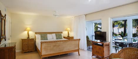 A hotel room with a bed, bedside tables, a television, a dining area with a table and chairs, and a balcony with a view of palm trees.