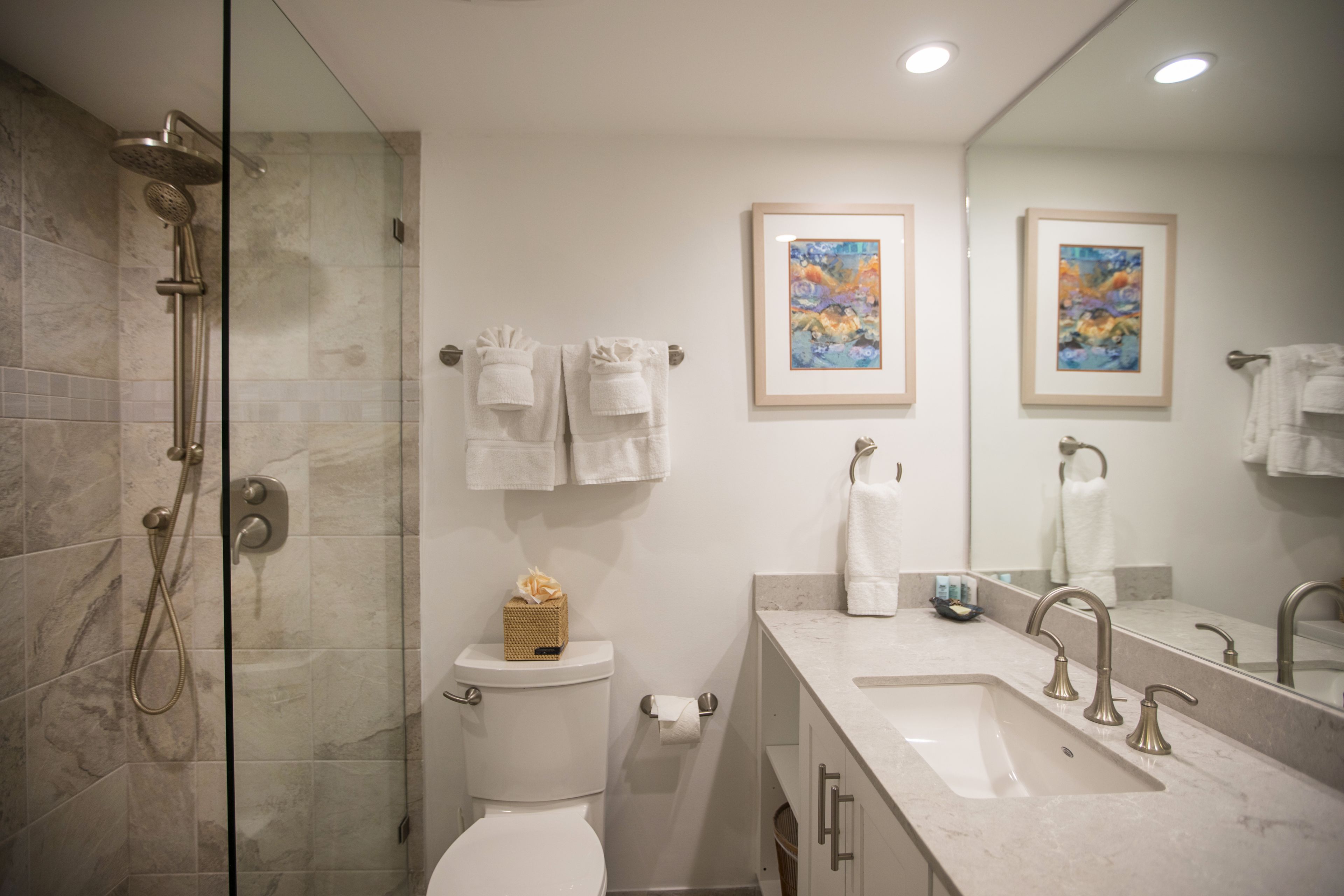 A modern bathroom with a glass-enclosed shower, a toilet, a sink with a marble countertop, and two framed artworks on the wall.