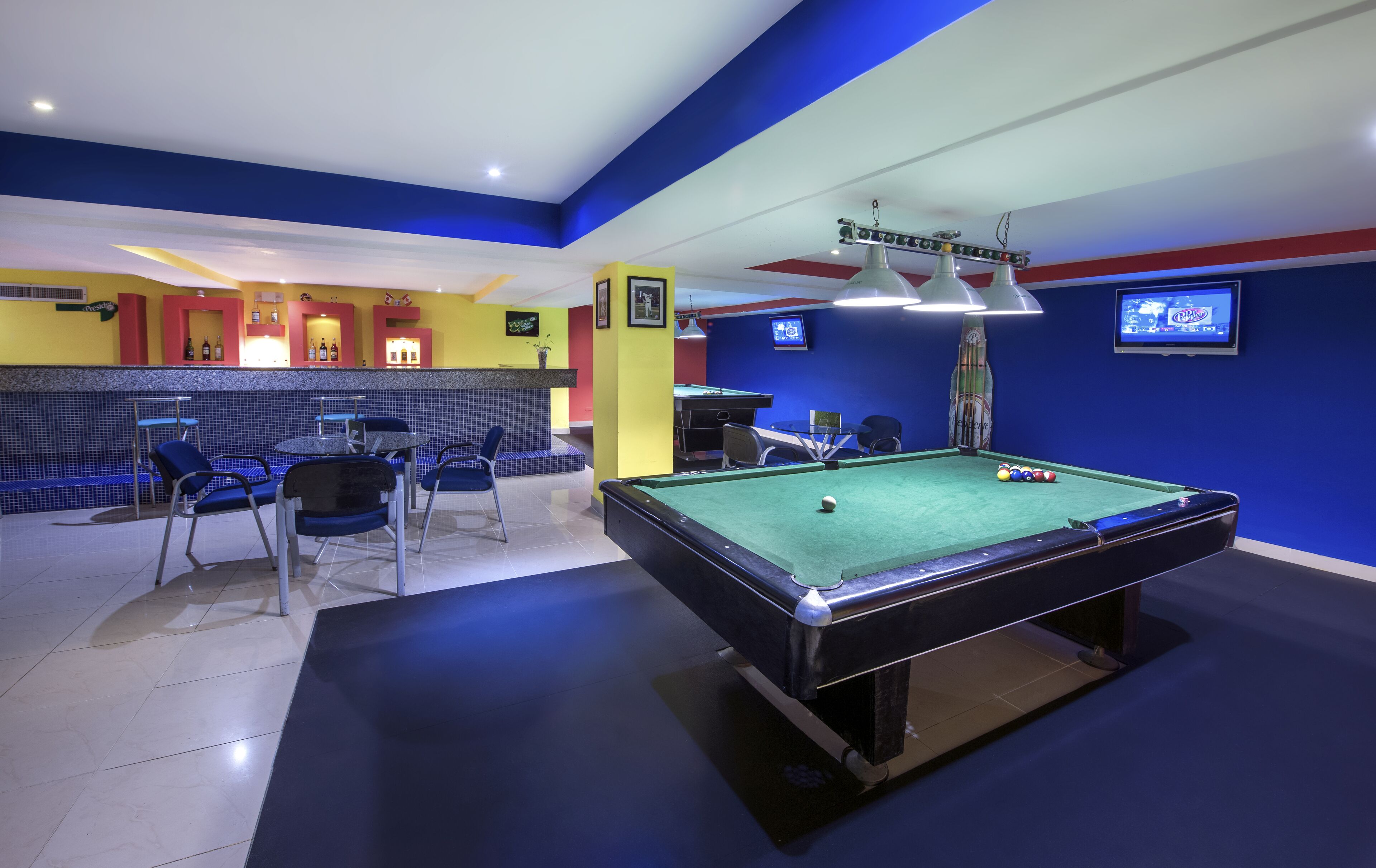 A pool table with chairs, a bar area, and a television screen in a room with blue and yellow walls.