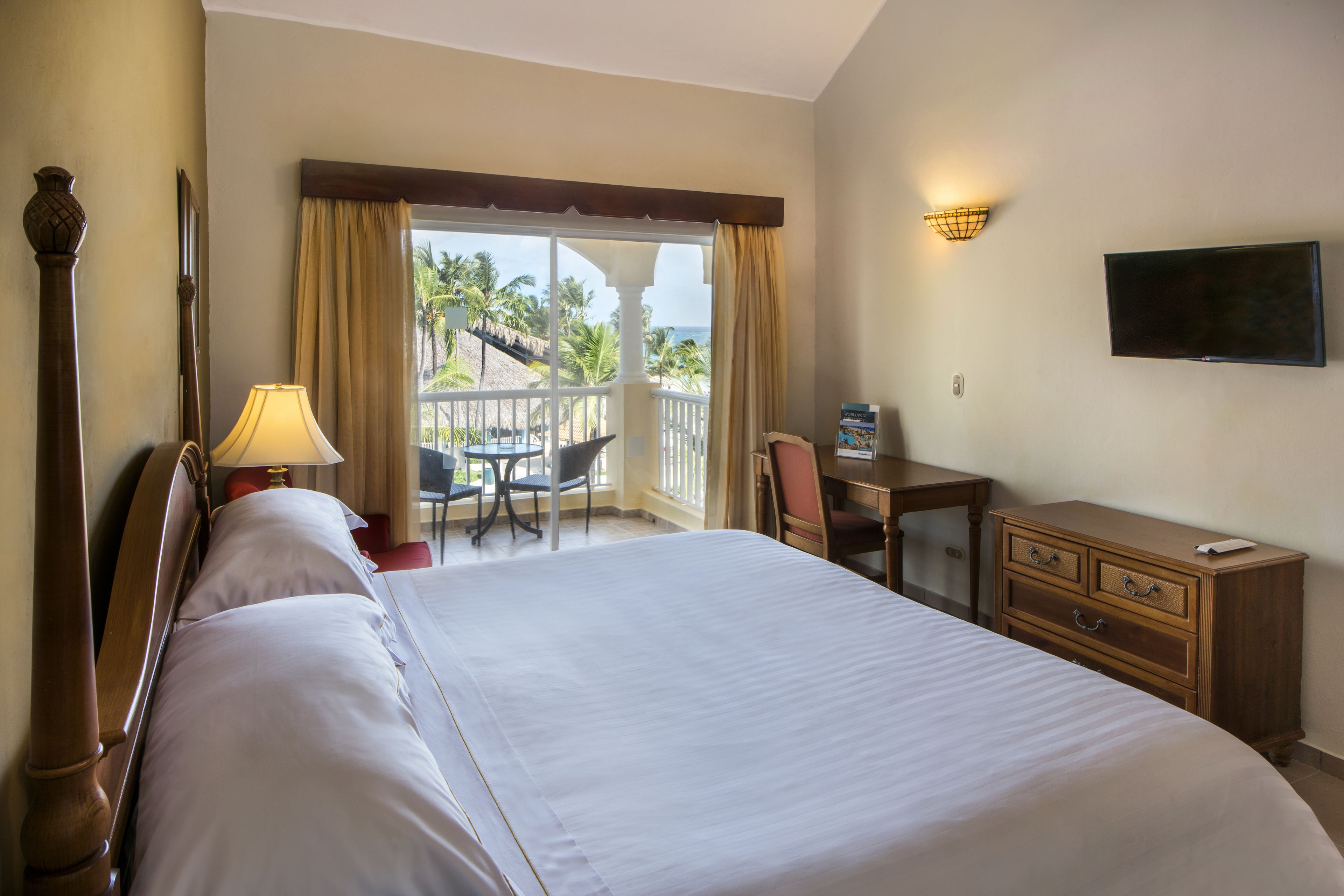 A hotel room with a large bed, a desk with a chair, a television, and a balcony with a view of palm trees and the ocean.