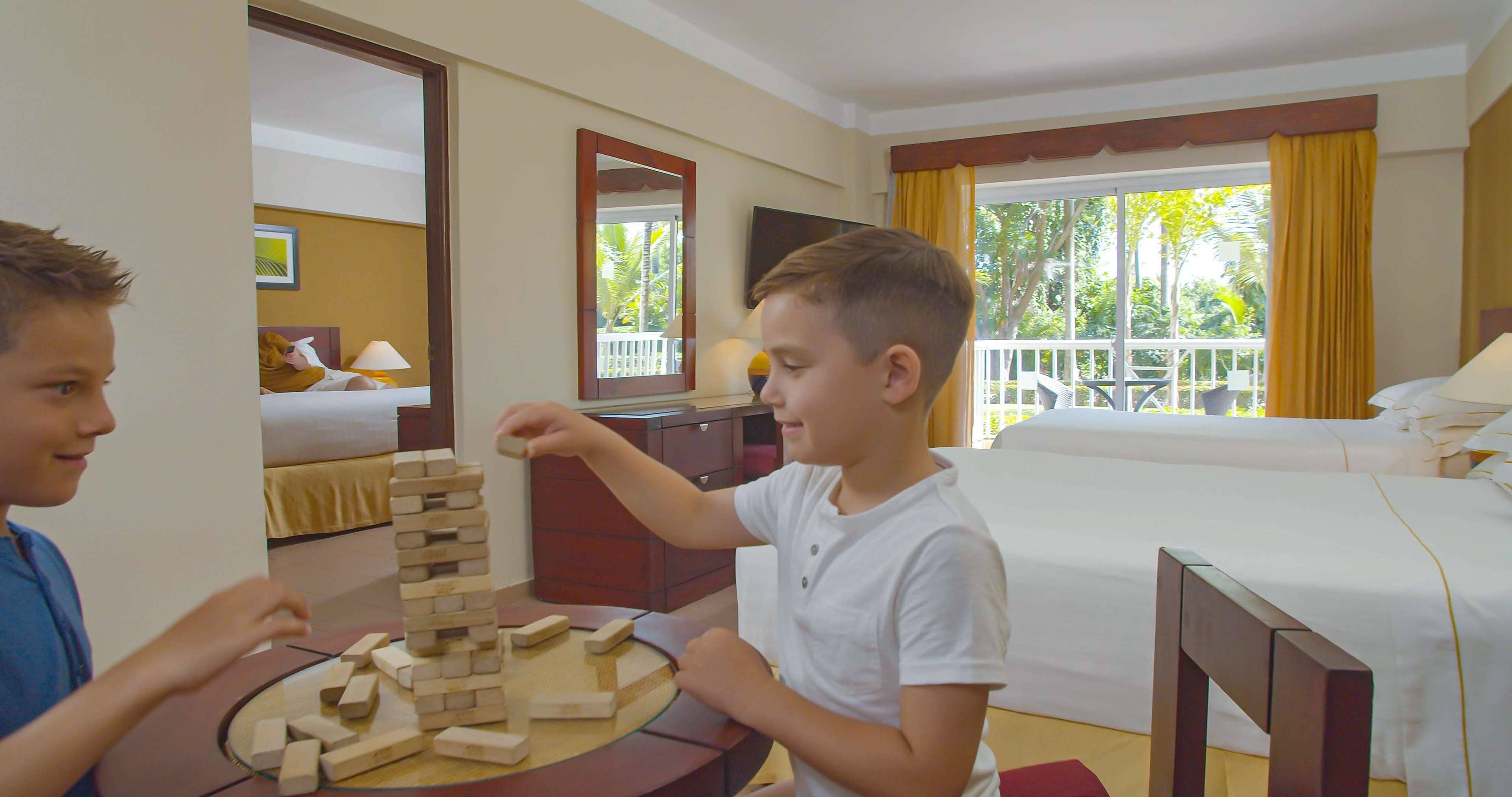 A hotel room with a bed, a wooden table with a jigsaw puzzle, a mirror, and a balcony with yellow curtains.