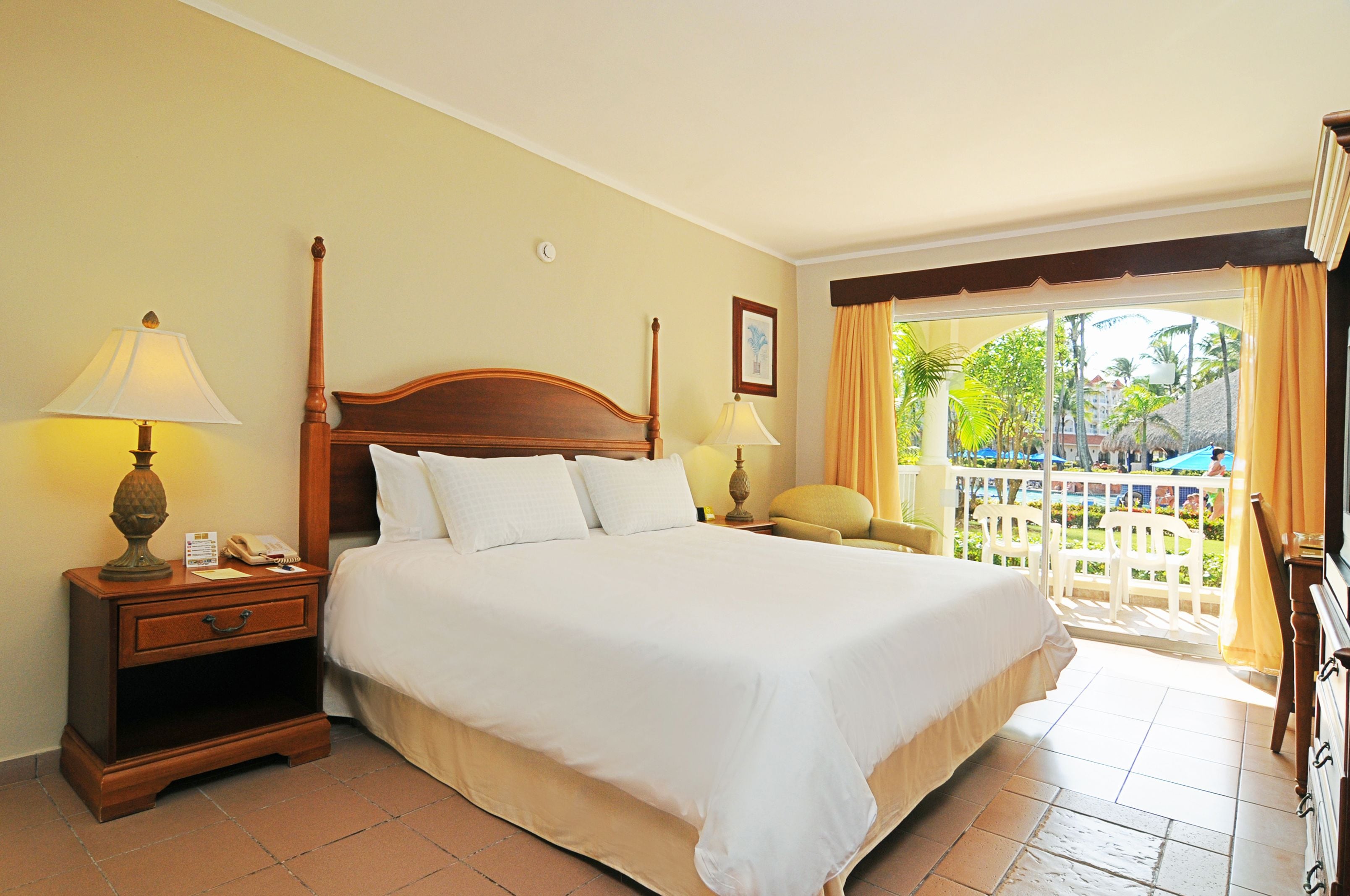 A hotel room with a large bed, two bedside tables with lamps, a wooden headboard, and a view of a pool and palm trees through a glass door.