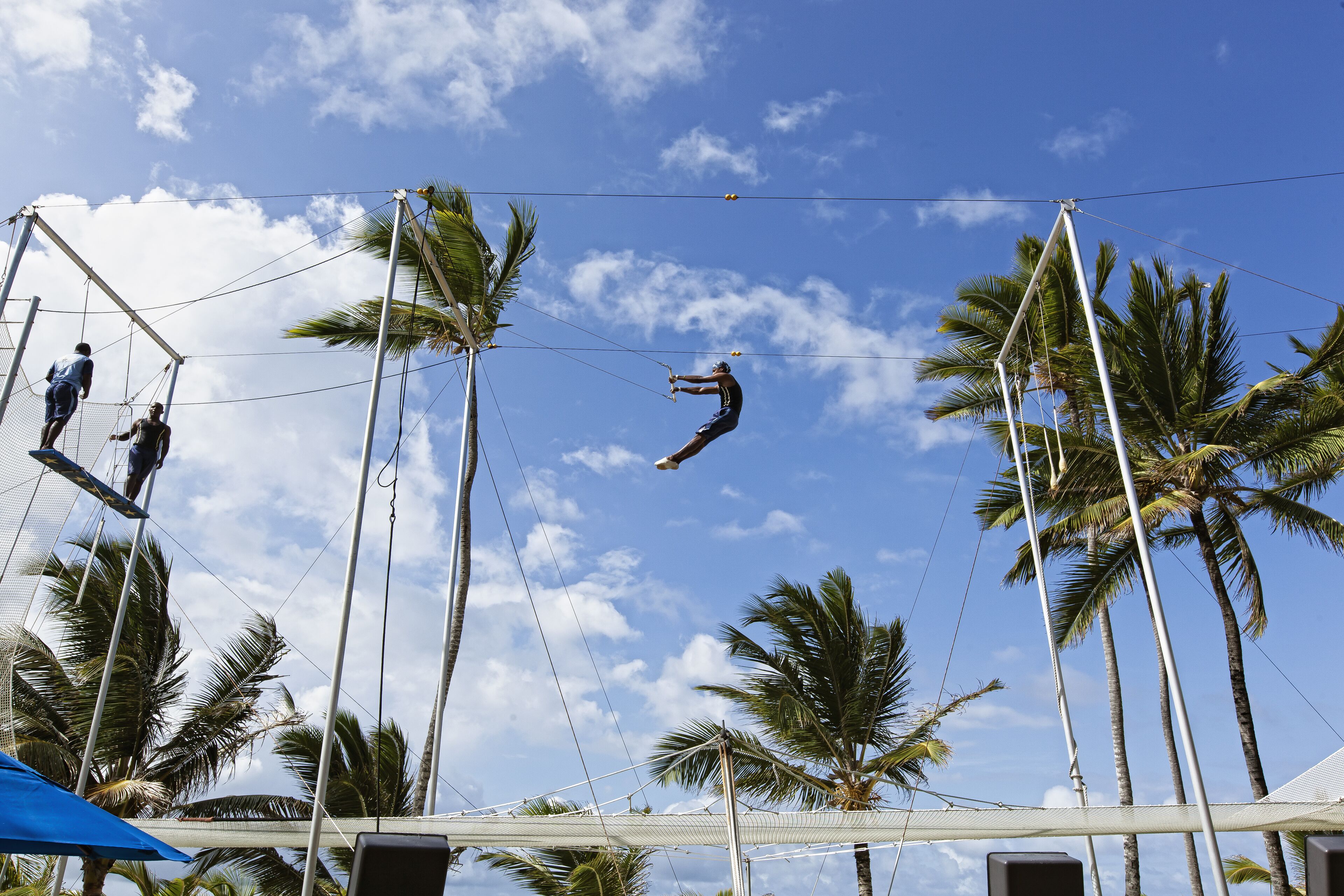 A trapeze artist performing in an outdoor setting with palm trees and a clear sky.