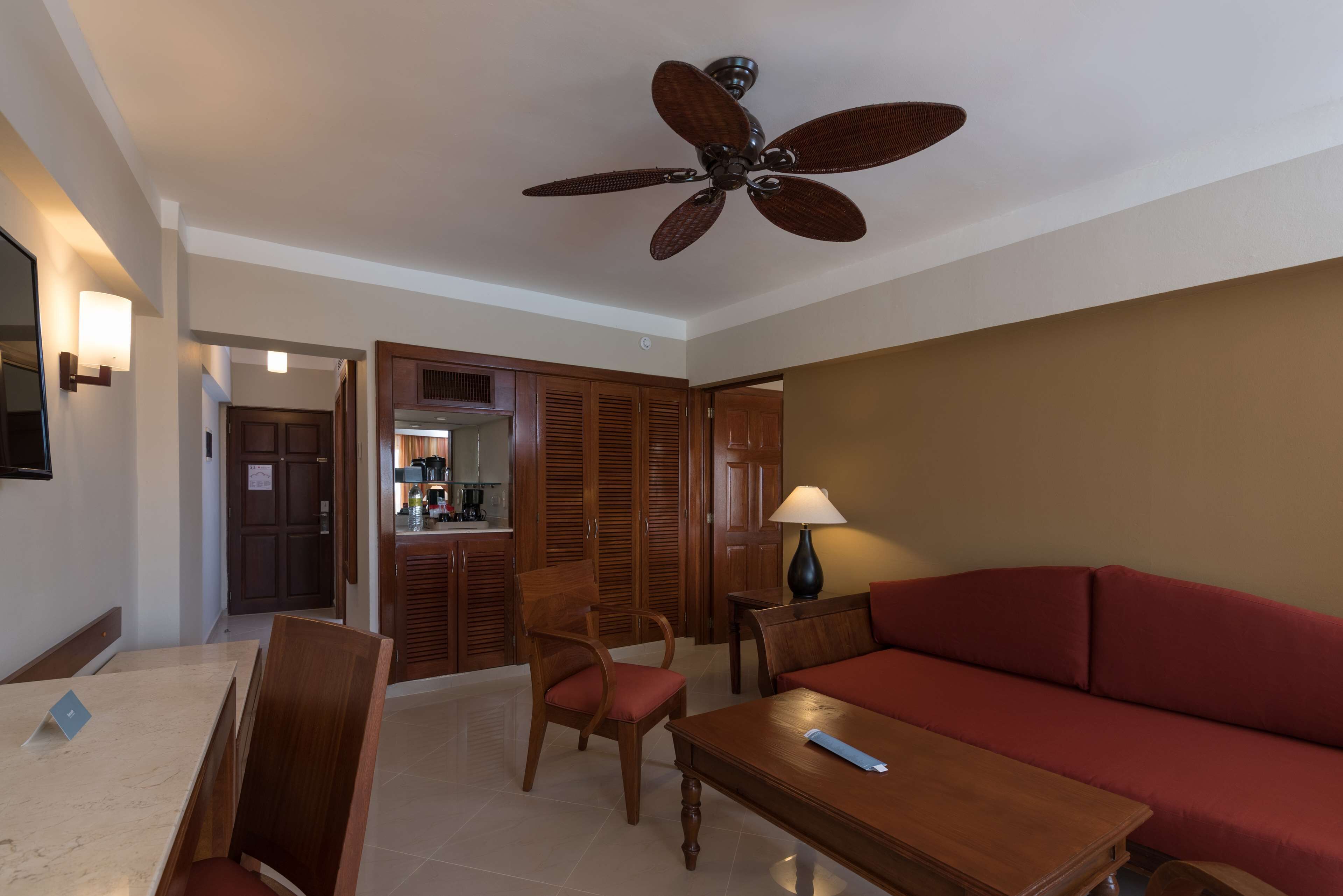 A hotel room with a wooden sofa, a dining table with chairs, a ceiling fan, and a kitchen area visible through an open door.