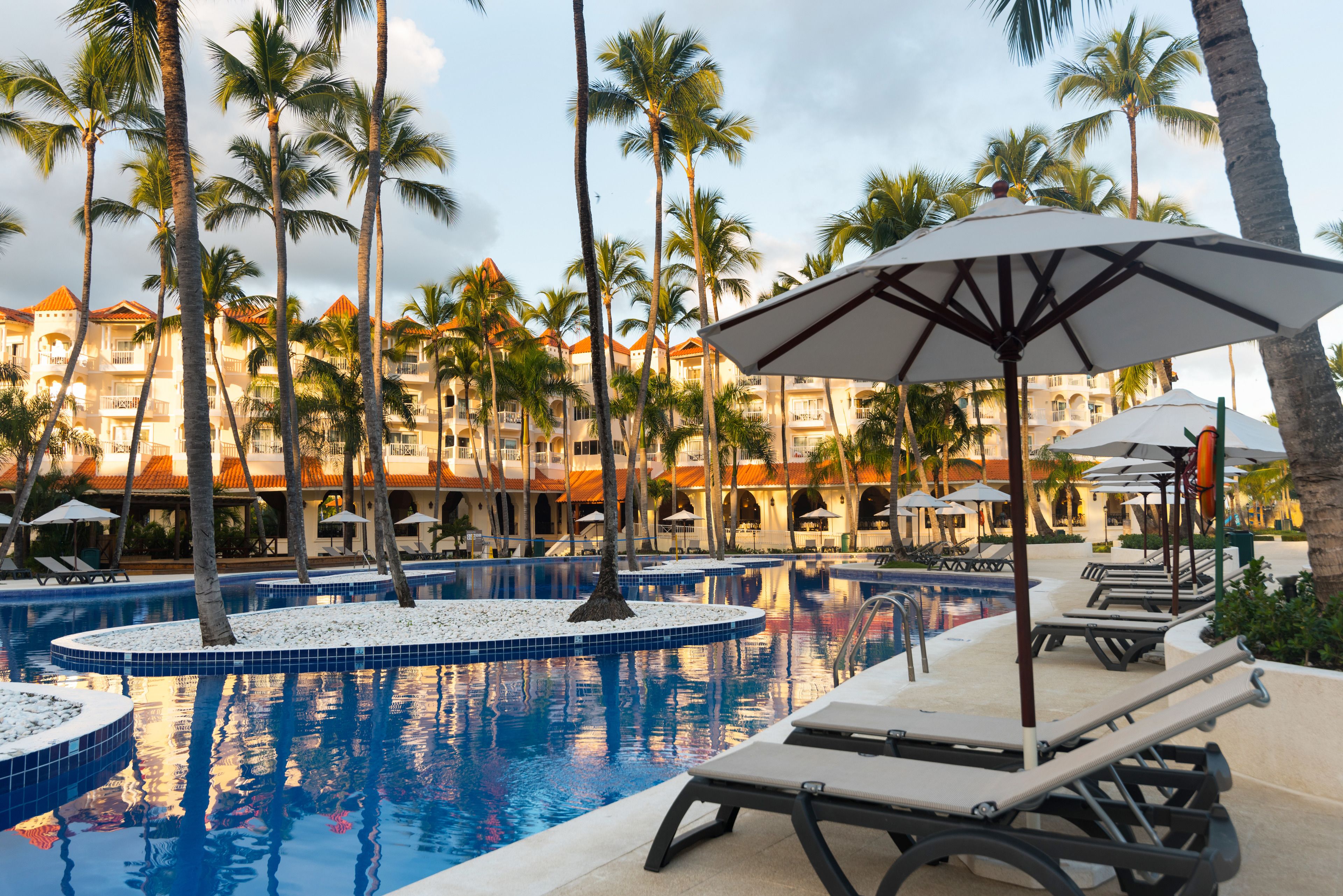 A pool area with lounge chairs and umbrellas, surrounded by palm trees and a building with balconies.