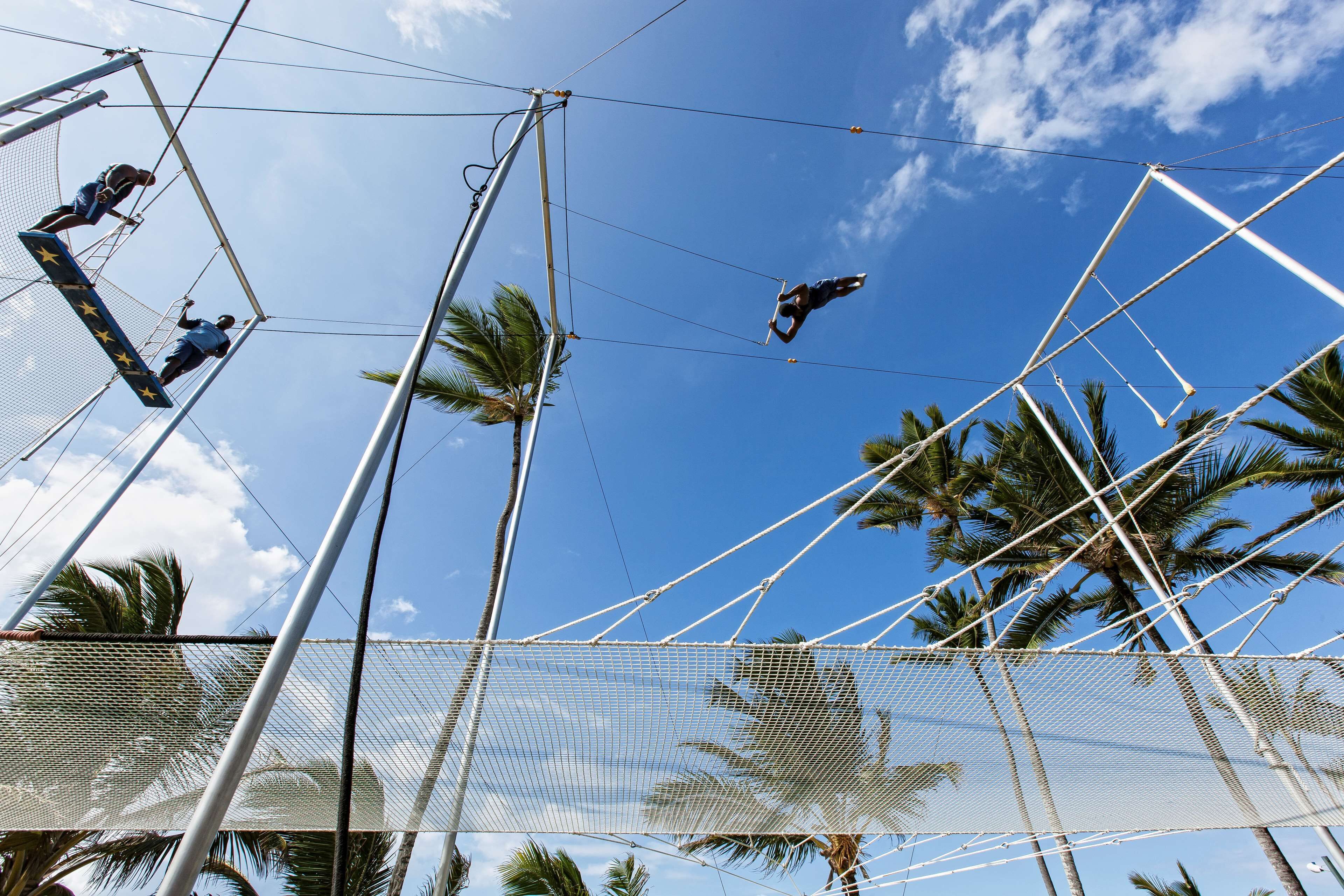 Un acto de trapecio con artistas, un cielo azul claro y palmeras al fondo.
