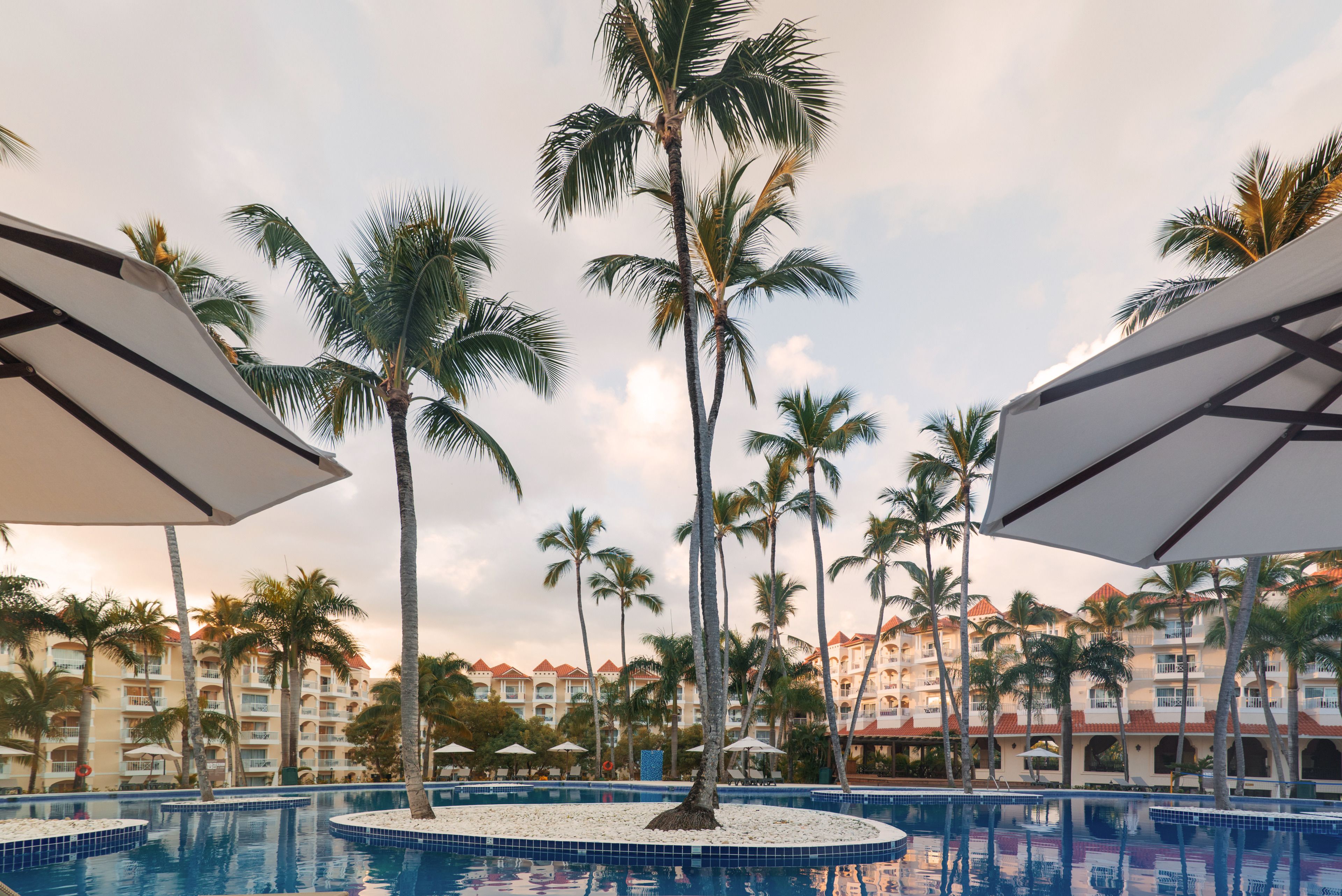 A pool area with palm trees and umbrellas, surrounded by hotel buildings.