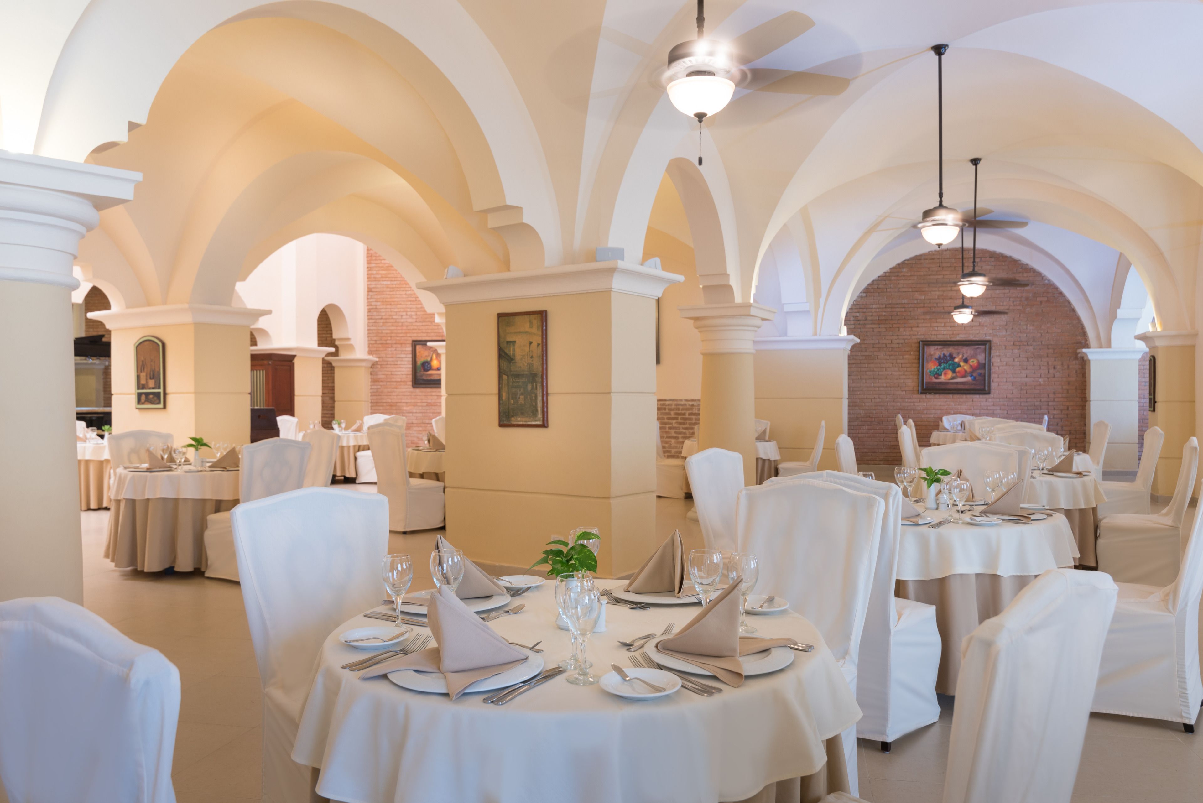 A restaurant with round tables set for dining, white chairs, and a brick wall in the background.