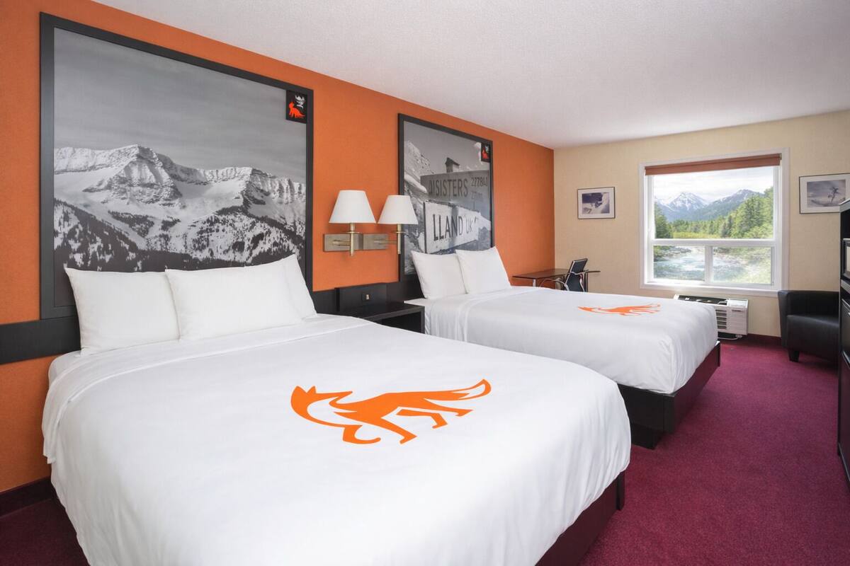A hotel room with two beds, each with a mountain landscape poster and a logo on the bedspread. There is a window with a mountain view, a desk with a chair, and a TV on a stand.