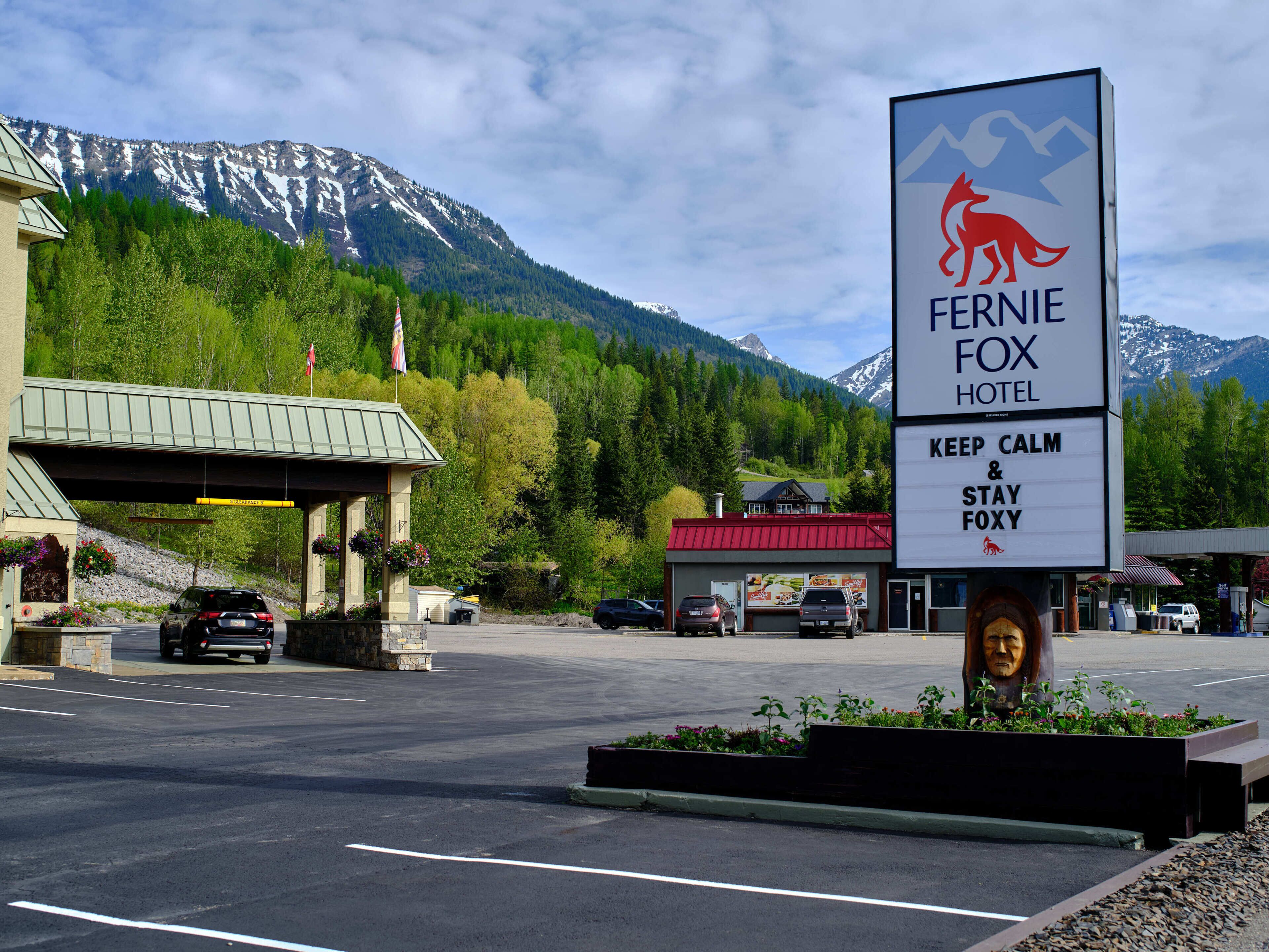 A hotel sign with a mountain backdrop, a parking area, and a building with a red roof.
