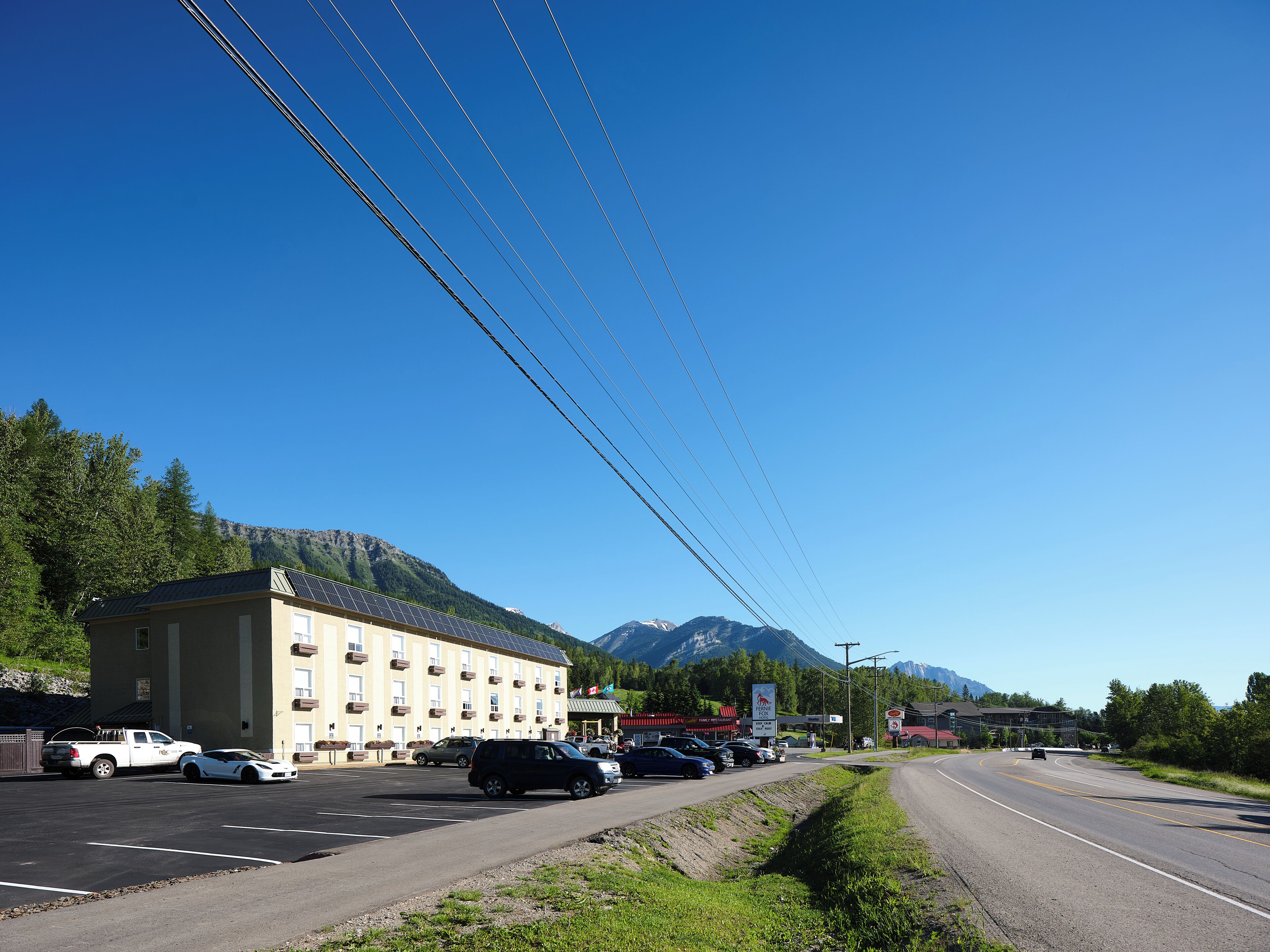 A building with a flat roof and multiple windows, adjacent to a road with parked cars and a mountain in the background.