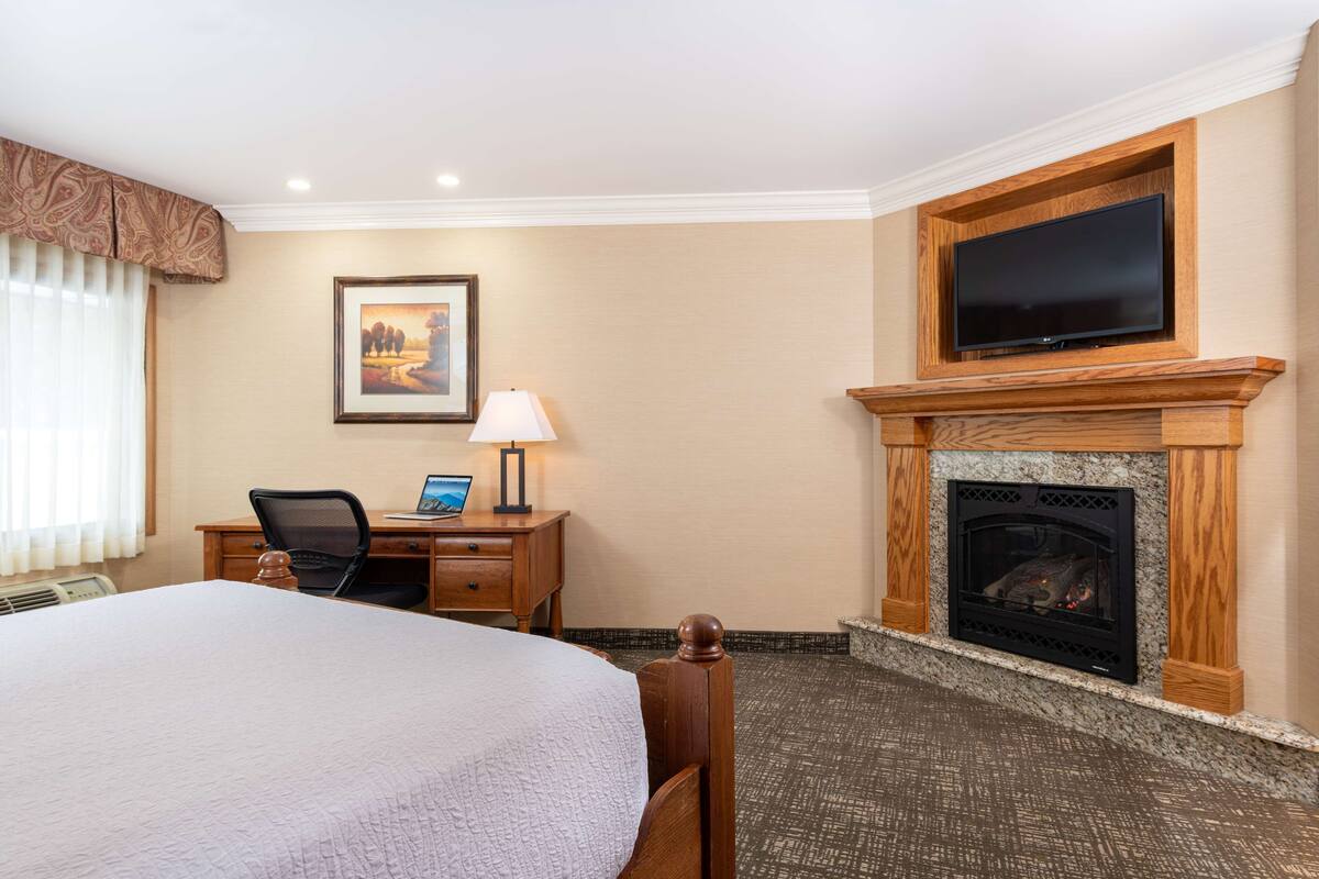 Une chambre d’hôtel avec un grand lit, un bureau avec un ordinateur portable, un téléviseur fixé au-dessus d’une cheminée et un tableau encadré au mur.