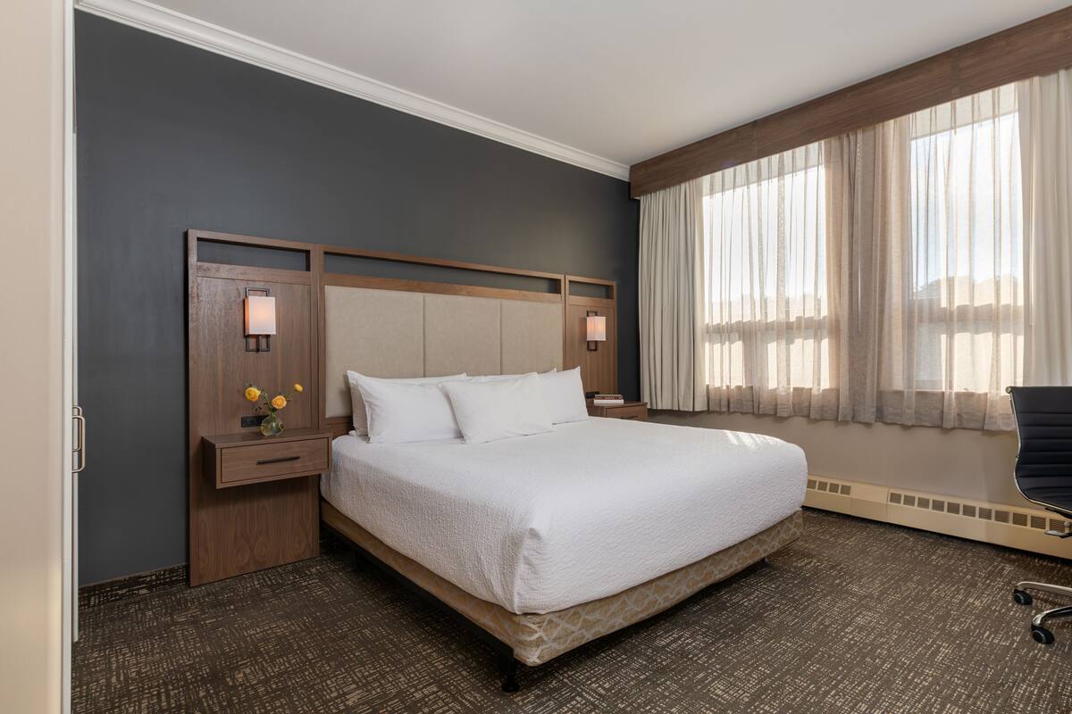 Une chambre d’hôtel avec un grand lit, une tête de lit en bois, deux lampes de chevet, une table de chevet, une chaise et une fenêtre avec des rideaux légers.