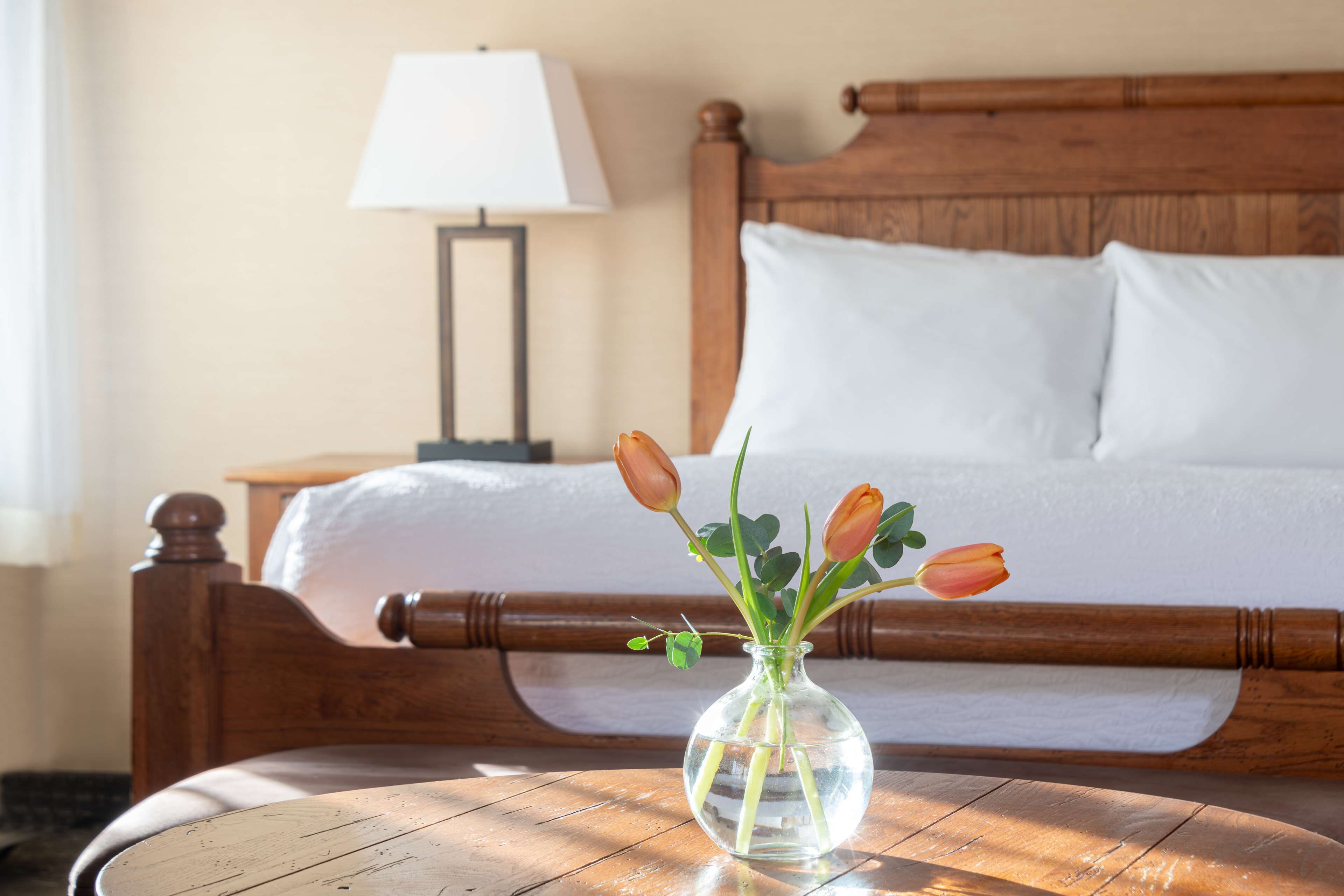 Una cama de madera con ropa de cama blanca, una mesita de noche con una lámpara, y un jarrón con flores sobre una mesa de madera.