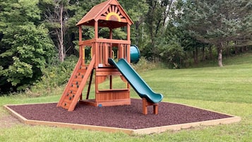 A wooden playground with a slide and climbing area, surrounded by a grassy field and trees.