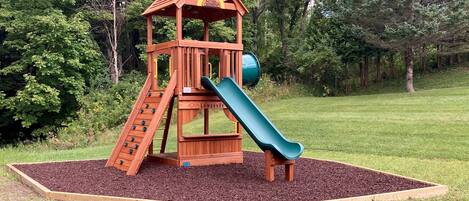 A wooden playground with a slide and climbing area, surrounded by a grassy field and trees.