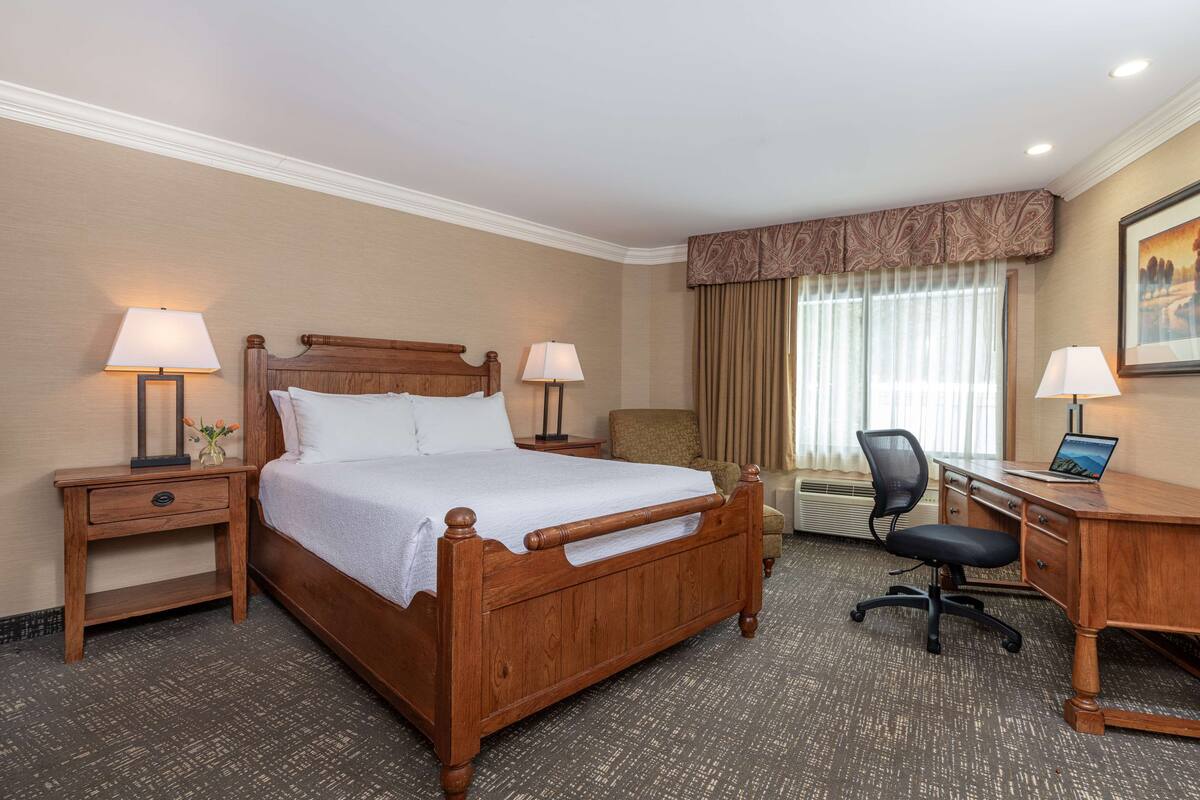 Habitación de hotel con una cama de madera, dos mesitas de noche con lámparas, un escritorio con silla y un ventanal con cortinas.