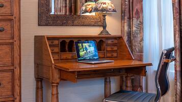 A wooden desk with a laptop, a lamp, and a chair in a room with patterned curtains and a mirror.