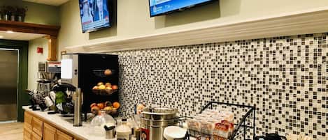 Cozinha de hotel com espaço para café, alimentos de pequeno-almoço e uma televisão a exibir um noticiário.
