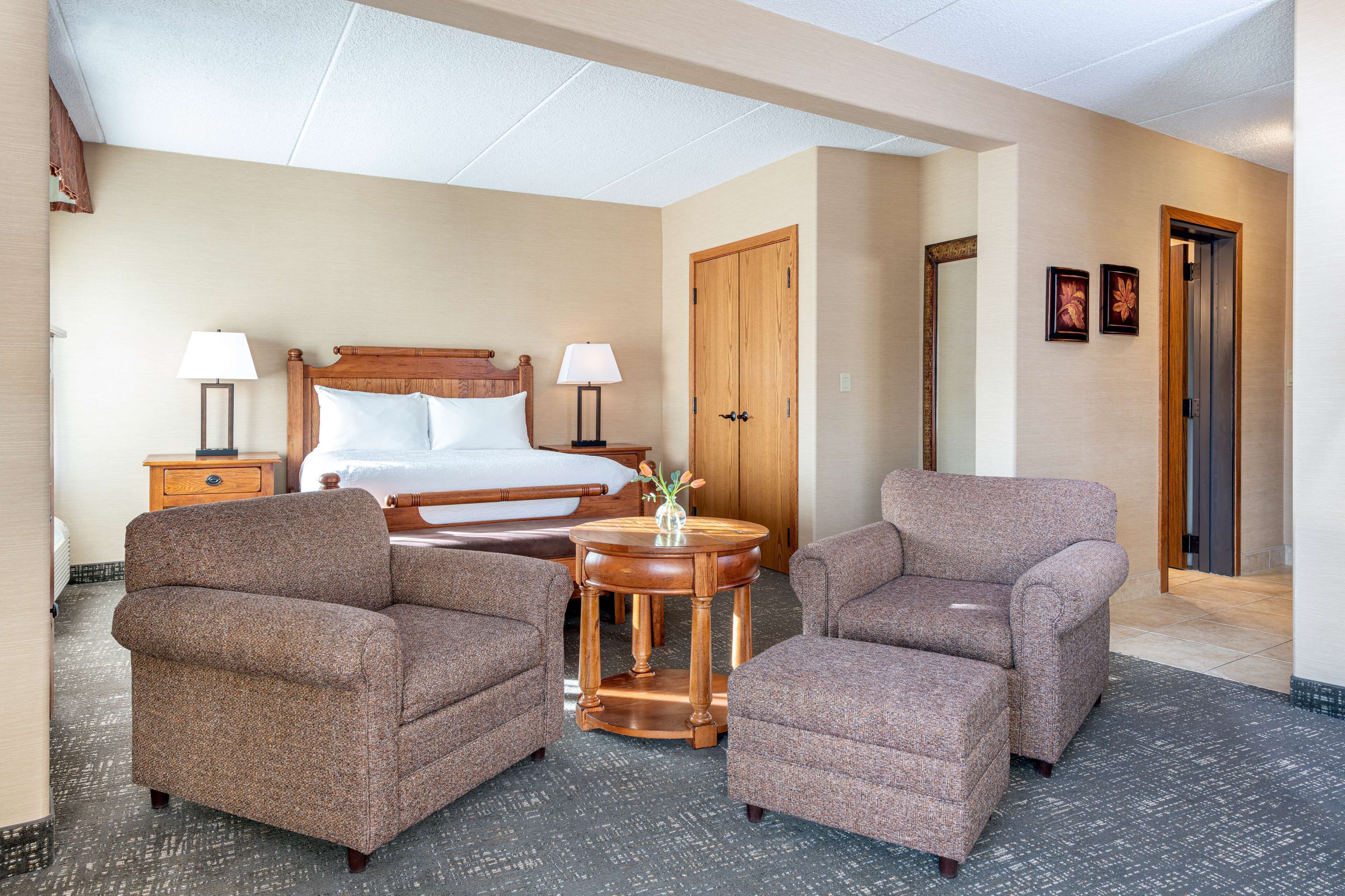 Une chambre d’hôtel comprenant un lit, deux fauteuils, une table ronde en bois avec un vase, et une table de chevet avec des lampes.