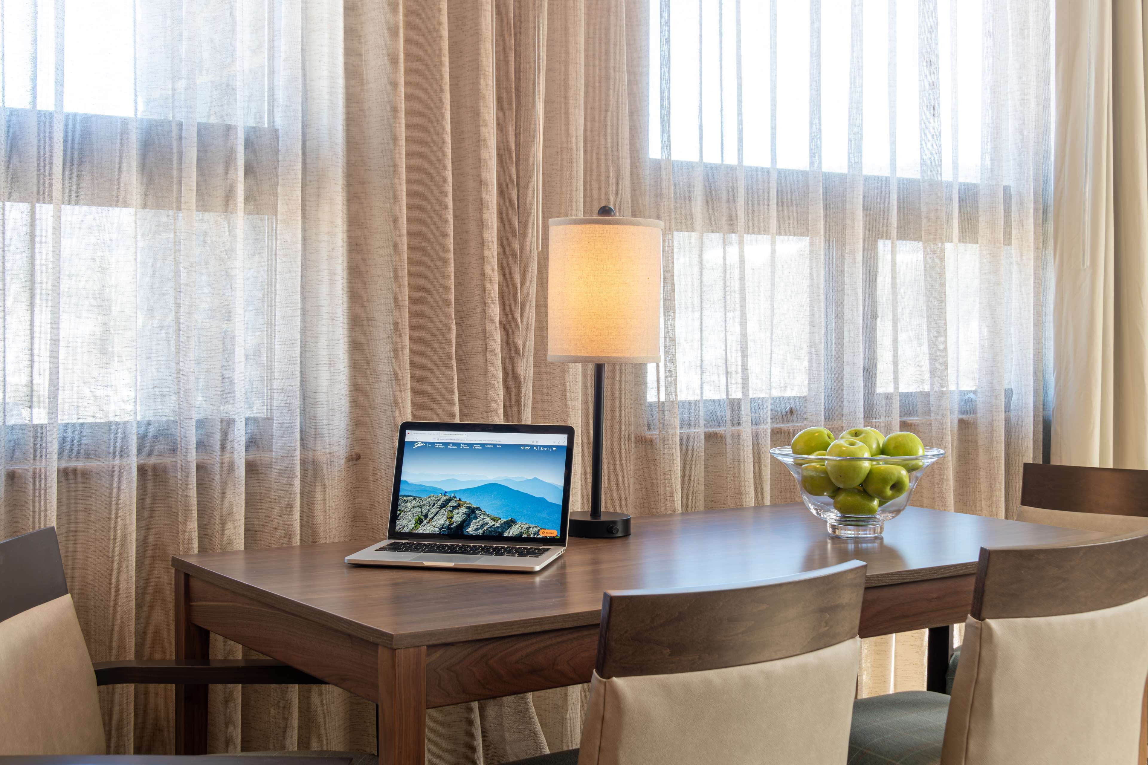 Habitación de hotel con un escritorio de madera, una computadora portátil, una lámpara y un bol de manzanas.