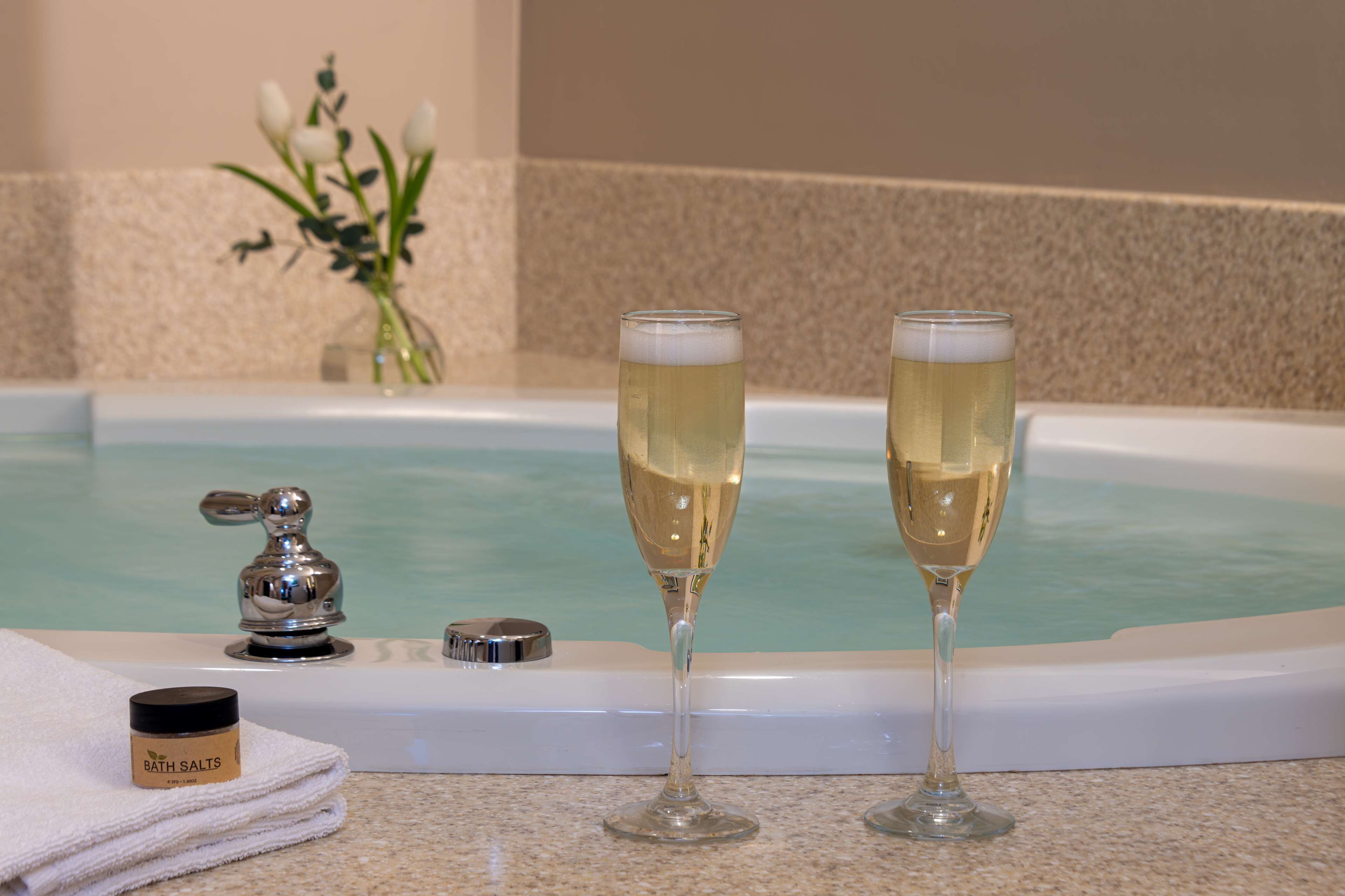 Two champagne glasses on a bathroom counter with a bathtub in the background.