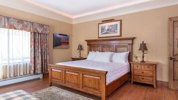 Un dormitorio con una cama grande de madera, una mesita de noche con una lámpara, otra mesita de noche más pequeña con una lámpara, un cuadro enmarcado sobre la cama y una ventana con cortinas.