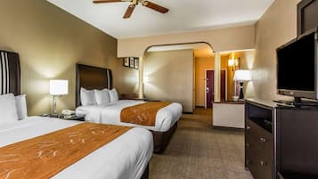 A hotel room with two beds, a desk with a TV, a ceiling fan, and a bathroom visible through an archway.