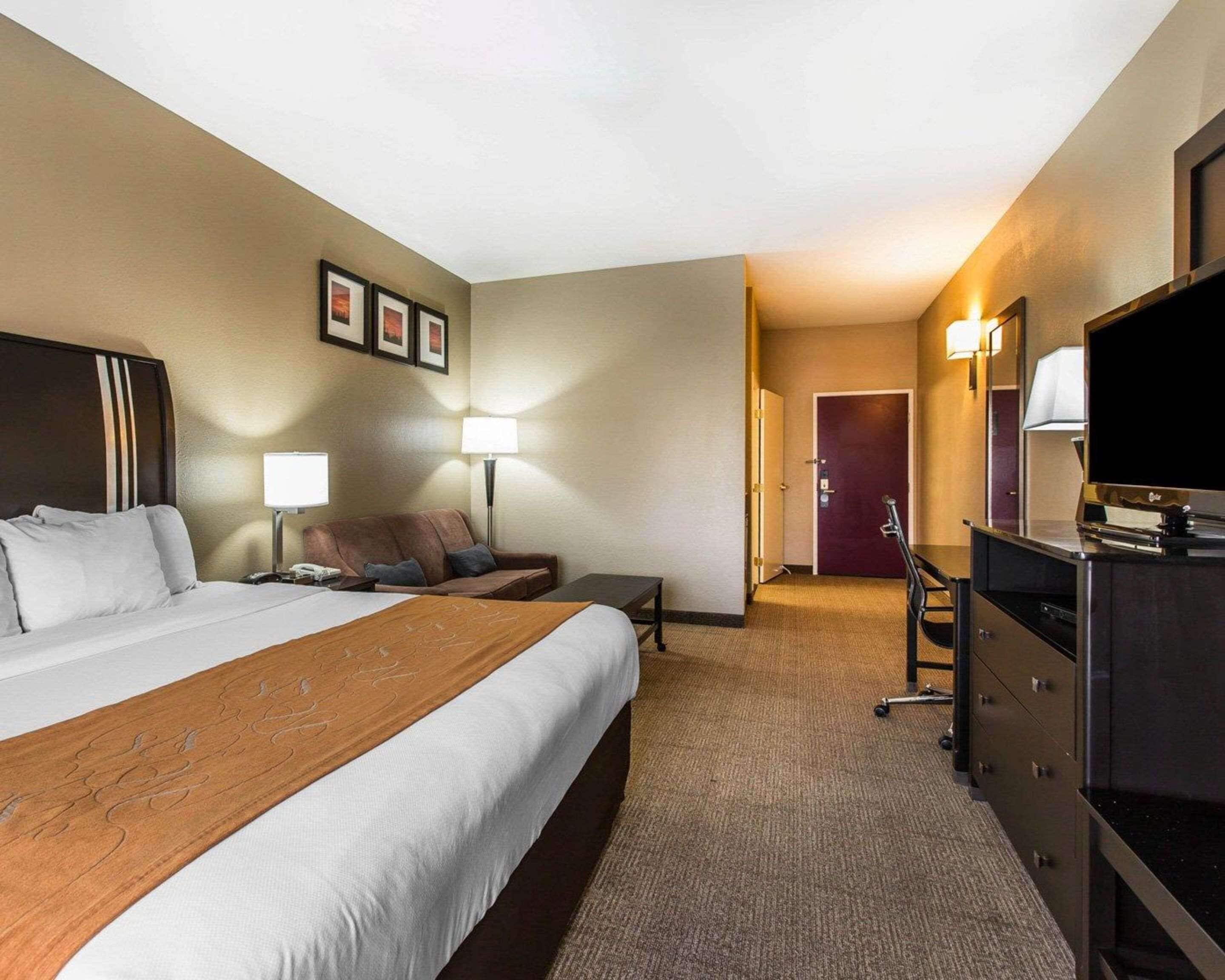 A hotel room with a large bed, a desk, a chair, a TV, and a door leading to another room.