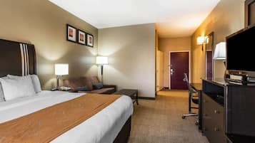 A hotel room with a large bed, a desk, a chair, a TV, and a door leading to another room.