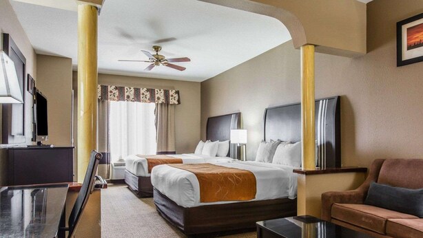 A hotel room with two beds, a desk, a chair, a sofa, a TV, and a ceiling fan.