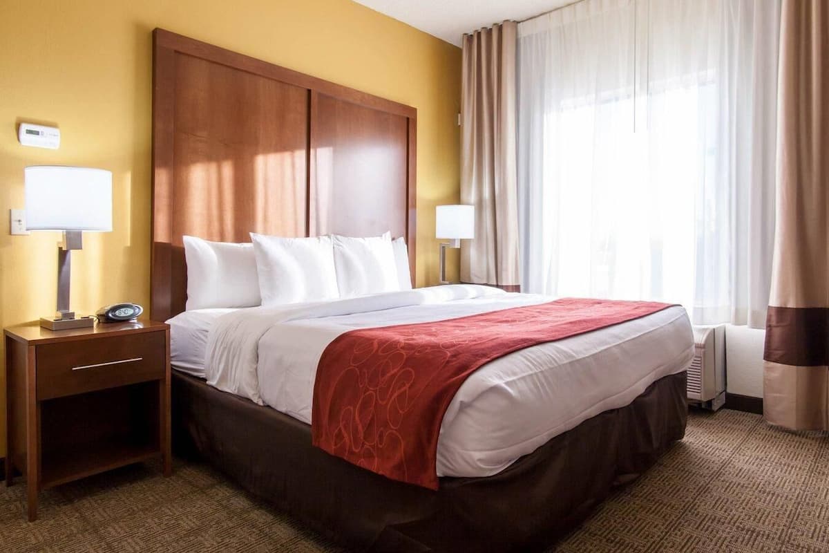 A hotel room with a large bed, two bedside tables, a nightstand, a red throw blanket, a wall clock, and a window with curtains.