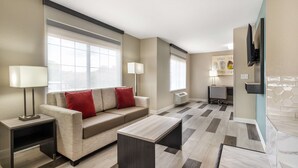 A modern living room with a sofa, coffee table, and side tables. There is a television mounted on the wall and a window with blinds. - Best Western Prime Inn & Suites (Poteau)