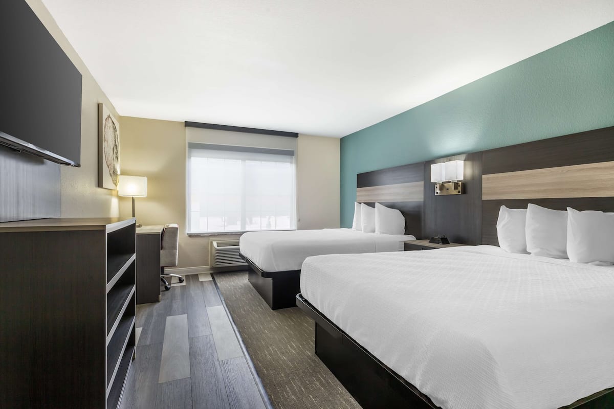 Quarto de hotel com duas camas, uma escrivaninha, uma cadeira, televisão e janela com persianas.