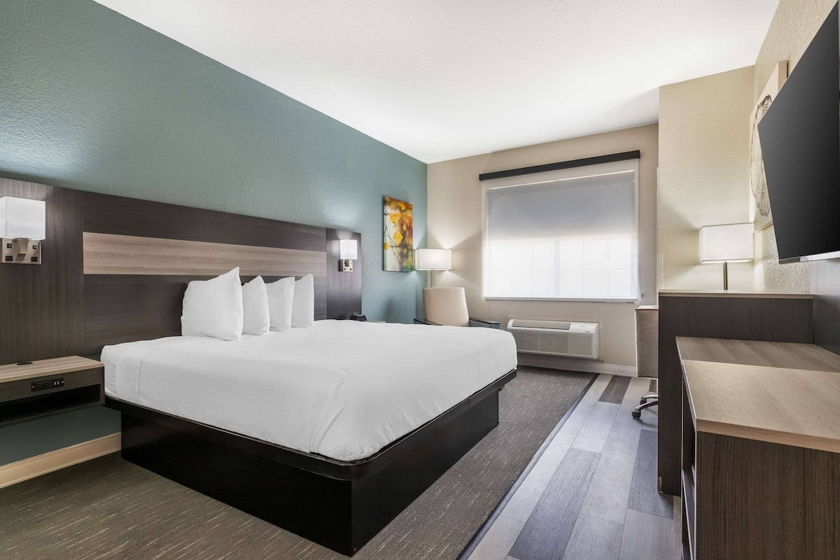 Quarto de hotel moderno com uma cama grande, mesas de cabeceira, escrivaninha e uma janela com persianas.