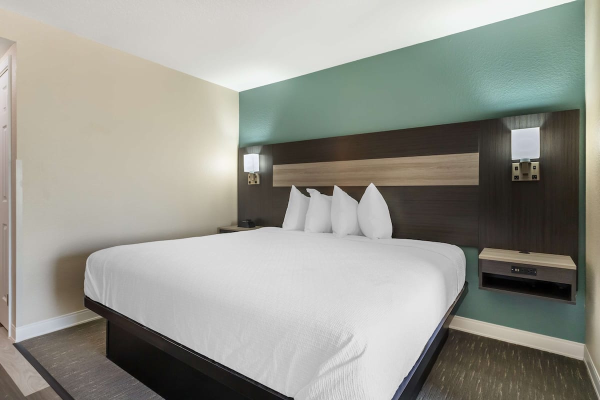 Quarto de hotel com uma cama grande, uma mesa de cabeceira, uma luminária na parede e um pequeno estante.