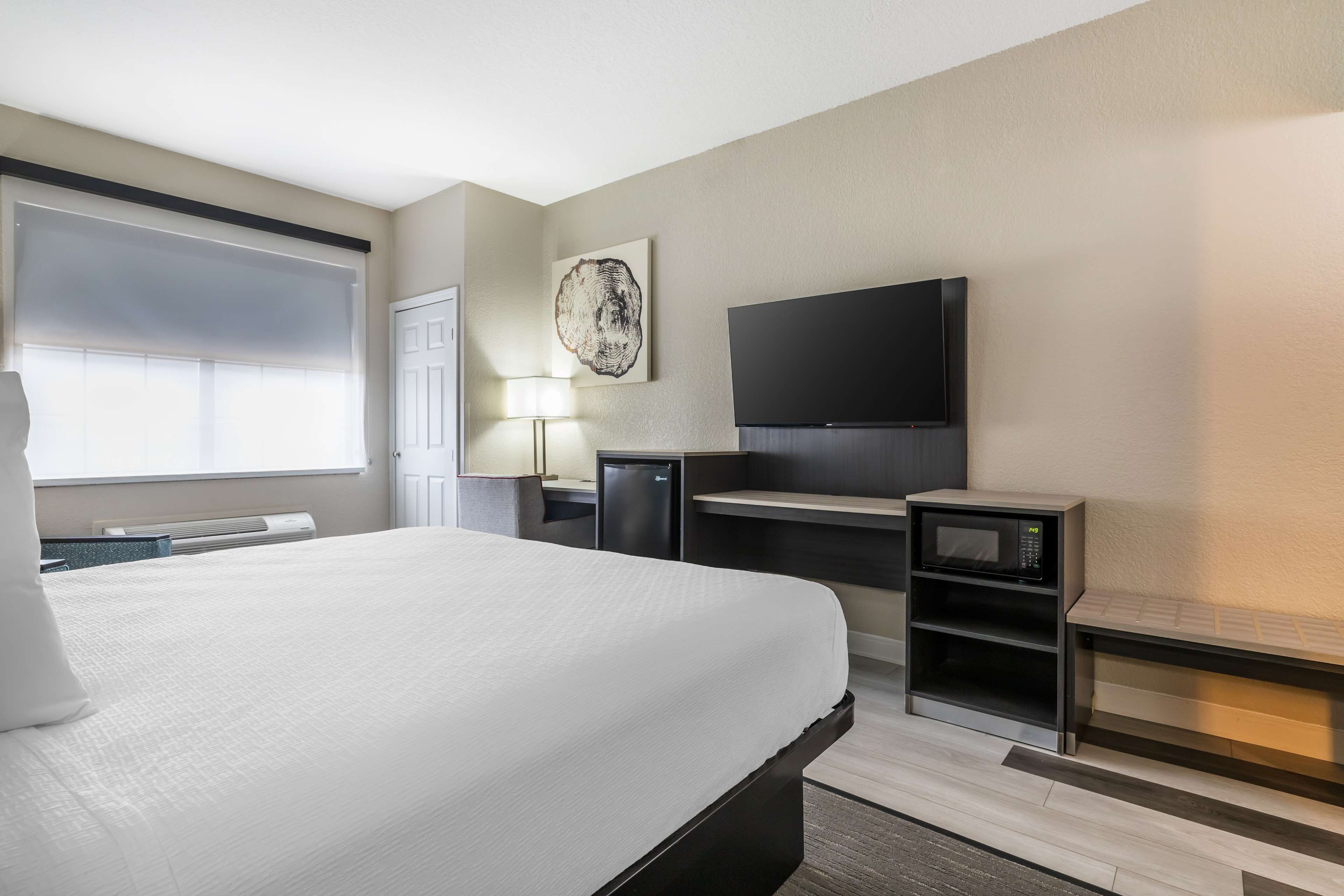 Ein Hotelzimmer mit einem großen Bett, einem flachbildfernseher an der Wand, einem Schreibtisch mit Lampe und einer Mikrowelle auf einem Regal.
