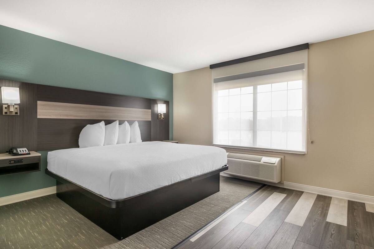 Quarto de hotel moderno com cama grande, mesa de cabeceira, luminária na parede e uma janela ampla com persianas.