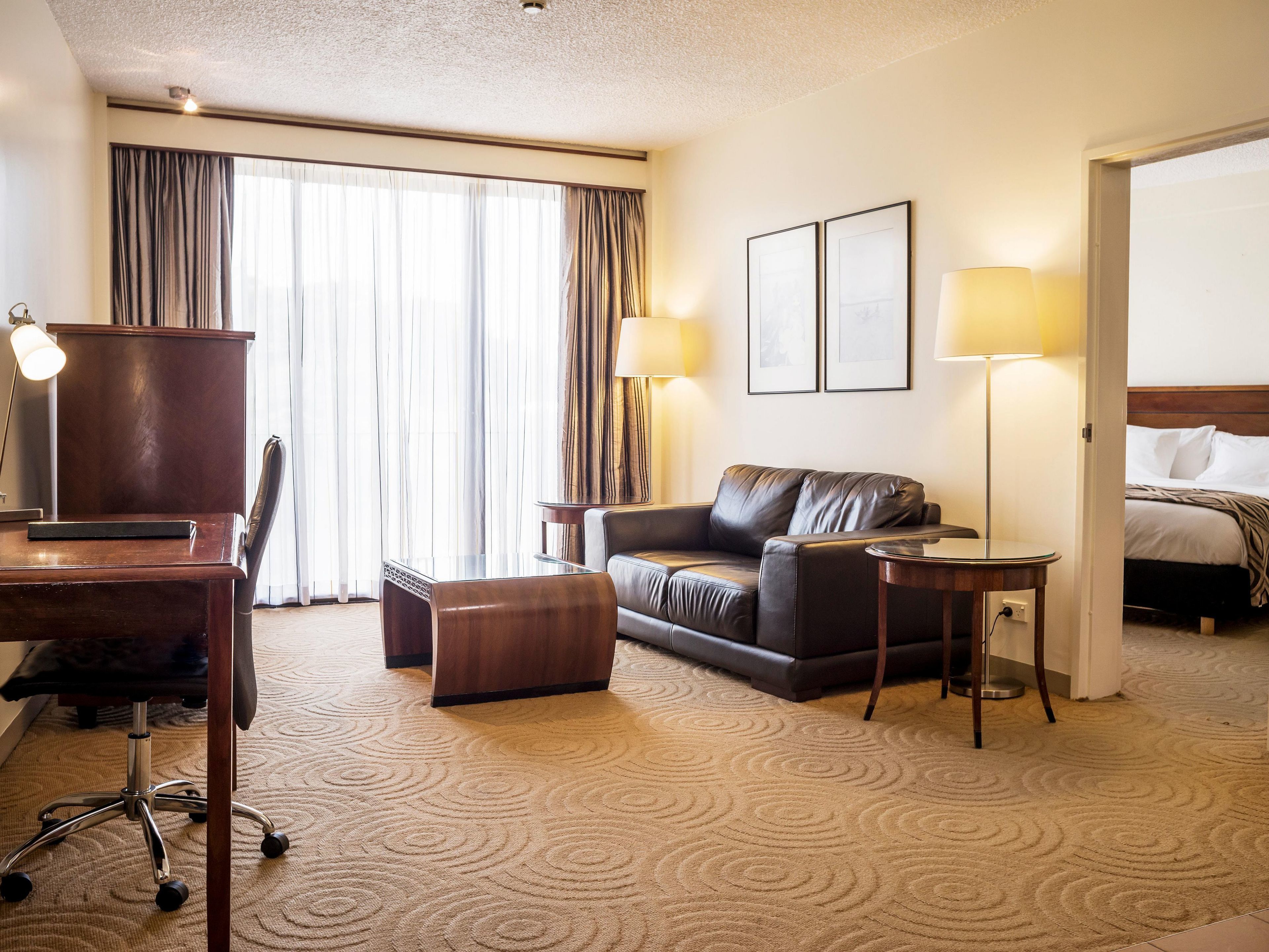 Habitación de hotel con sofá de cuero, un escritorio con una silla, cama, dos lámparas y un amplio ventanal con cortinas.