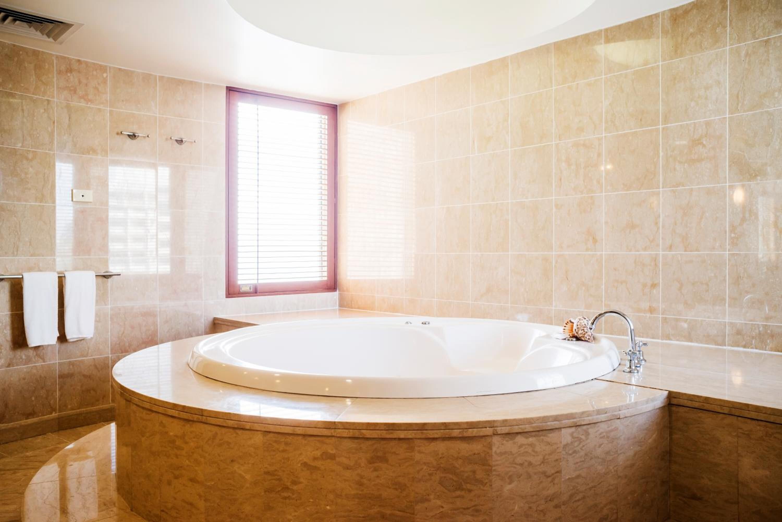 A modern bathroom with a large, round bathtub, a window with blinds, and a marble-tiled wall.