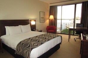 A hotel room with a large bed, a desk with a computer, a chair, a lamp, and a view of the water through large windows. - Crown Hotel (Port Moresby)