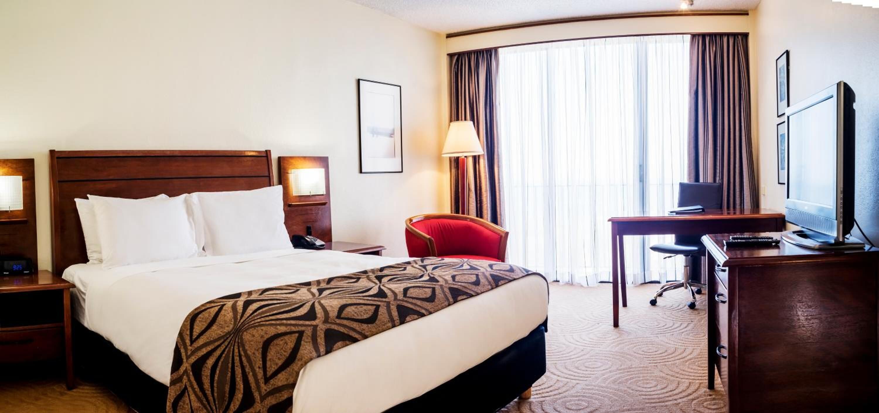 A hotel room with a large bed, a desk, a chair, a TV, and a window with curtains.