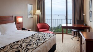 Une chambre d’hôtel équipée d’un lit, d’un bureau, d’une chaise, d’une lampe, d’une table de chevet et offrant une vue sur la mer grâce à de grandes fenêtres.