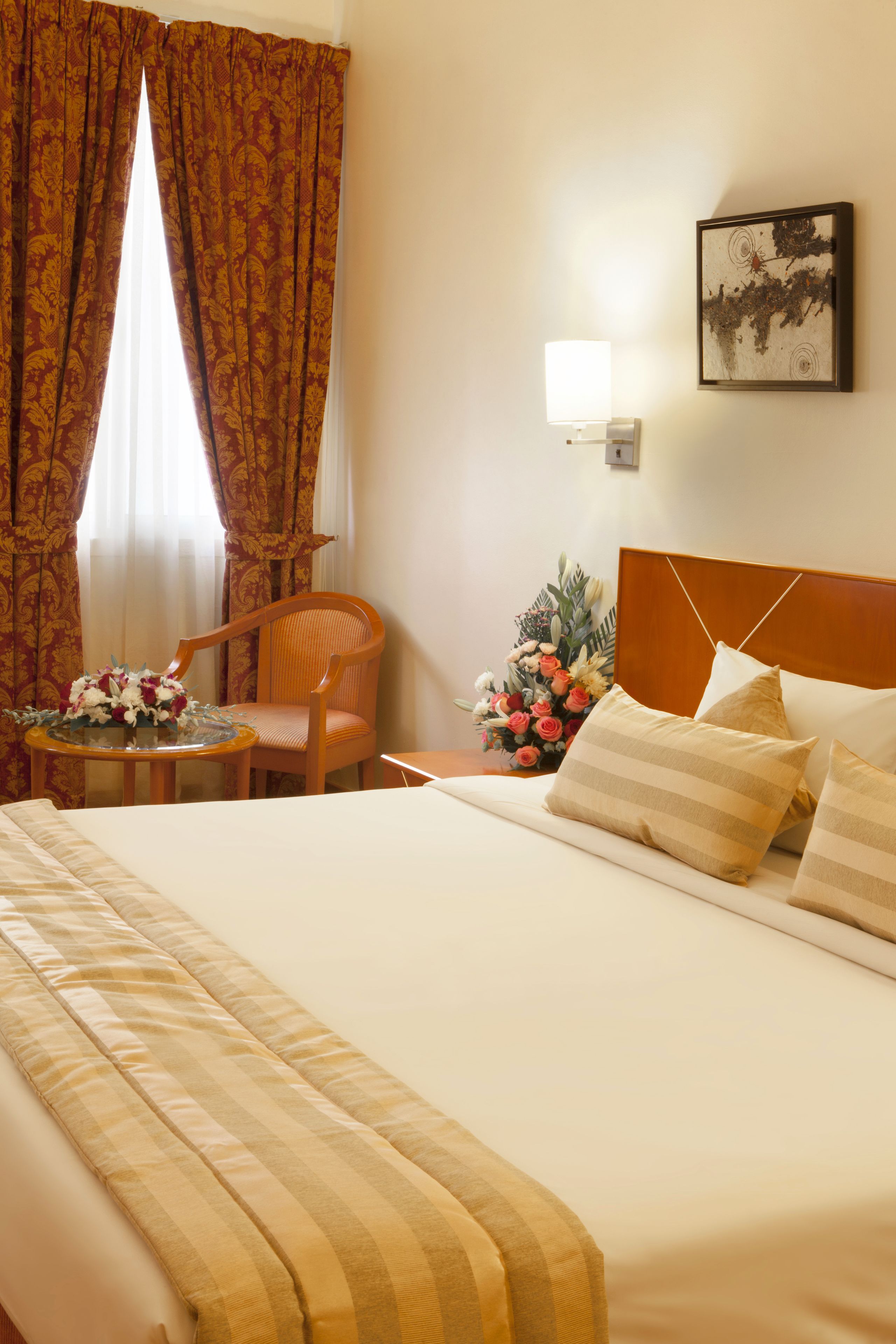 A hotel room with a large bed, a wooden chair, a small table with a vase of flowers, and a wall-mounted picture.
