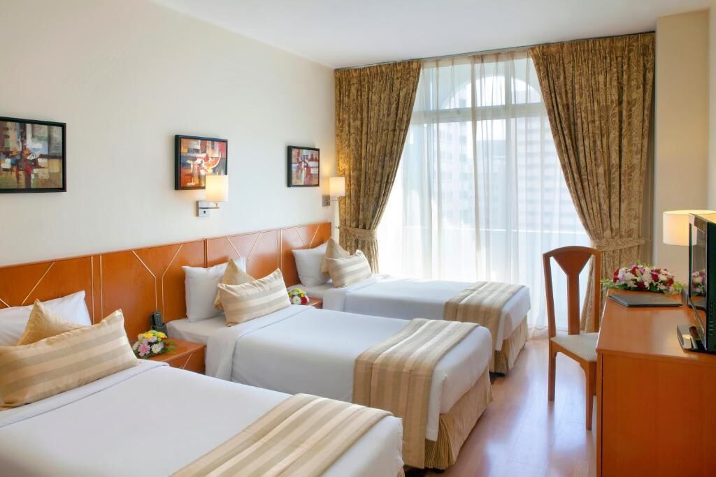 A hotel room with three beds, a wooden headboard, a desk with a chair, and a large window with curtains.