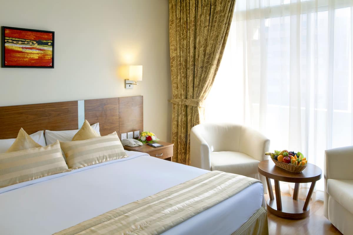 Habitación de hotel con una cama grande, una mesita de noche con teléfono y un jarrón de flores, una silla y una cesta de frutas sobre una mesita auxiliar.