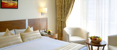 A hotel room with a large bed, a bedside table with a phone and a vase of flowers, a chair, and a fruit basket on a side table.