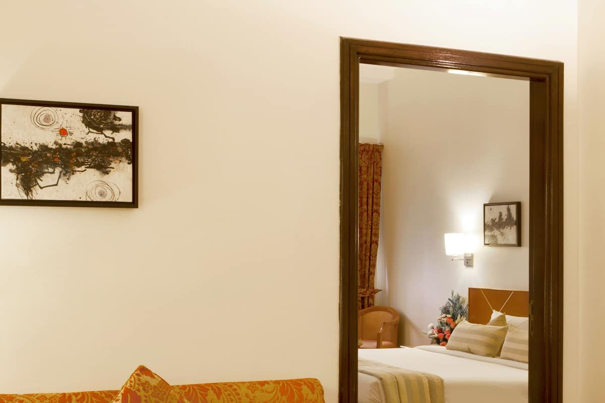 Habitación de hotel con sofá, una mesa de centro de cristal con fruta, una cama con cabecero y un espejo que refleja la habitación.