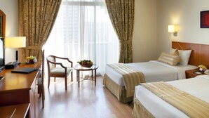 A hotel room with two beds, a desk, a chair, a small table with a fruit bowl, and a window with curtains. - Landmark Hotel (Dubai)